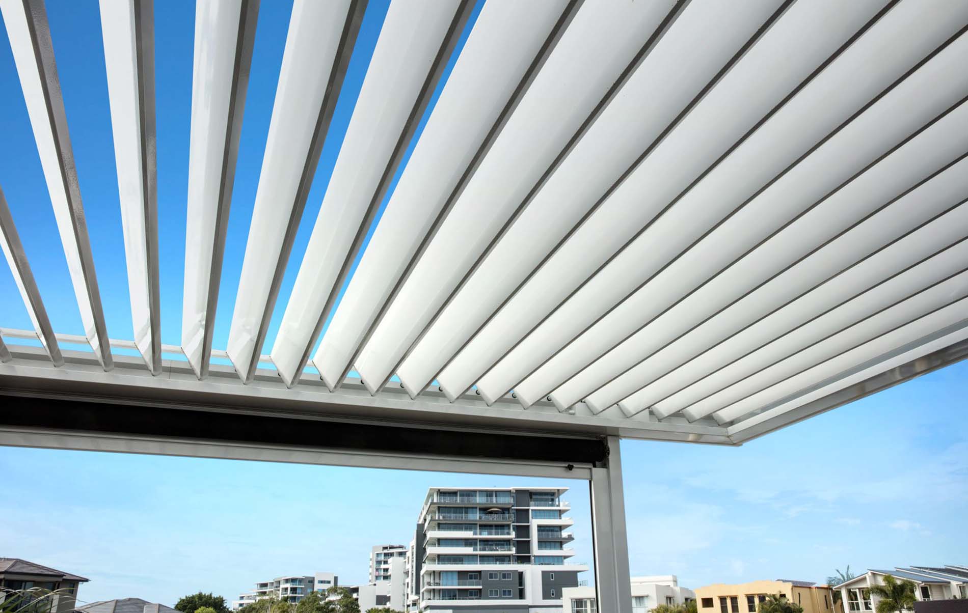 Adjustable white aluminium louvre roof slats over outdoor patio against clear blue sky with city skyline in background